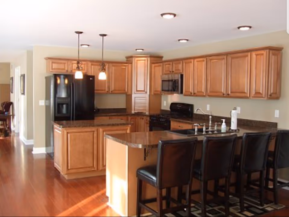 A spacious kitchen with wooden cabinets, a black refrigerator, a black stove with a microwave above it, and a kitchen island with a granite countertop. Four black leather bar stools are lined up along the island, and pendant lights hang above it. The floor is wooden, and the walls are painted beige.