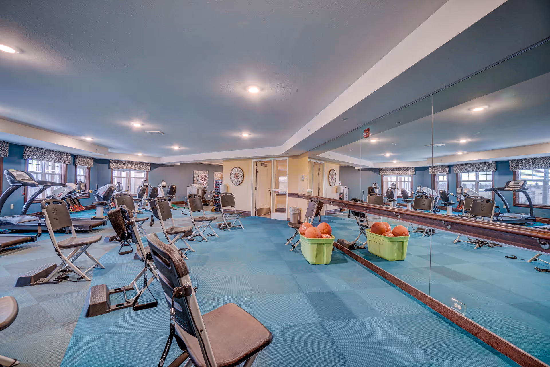 A spacious fitness room with blue carpeted floor, several exercise chairs arranged in rows, treadmills along the windows, and a large wall mirror reflecting the room. There are two green bins filled with orange exercise balls near the mirror. The room is well-lit with ceiling lights and has windows with blue valances.