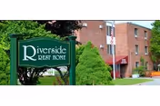 Exterior view of Riverside Rest Home, a brick building with multiple windows, surrounded by greenery including trees and bushes. A green sign with white text reading 'Riverside Rest Home' is prominently displayed in the foreground.
