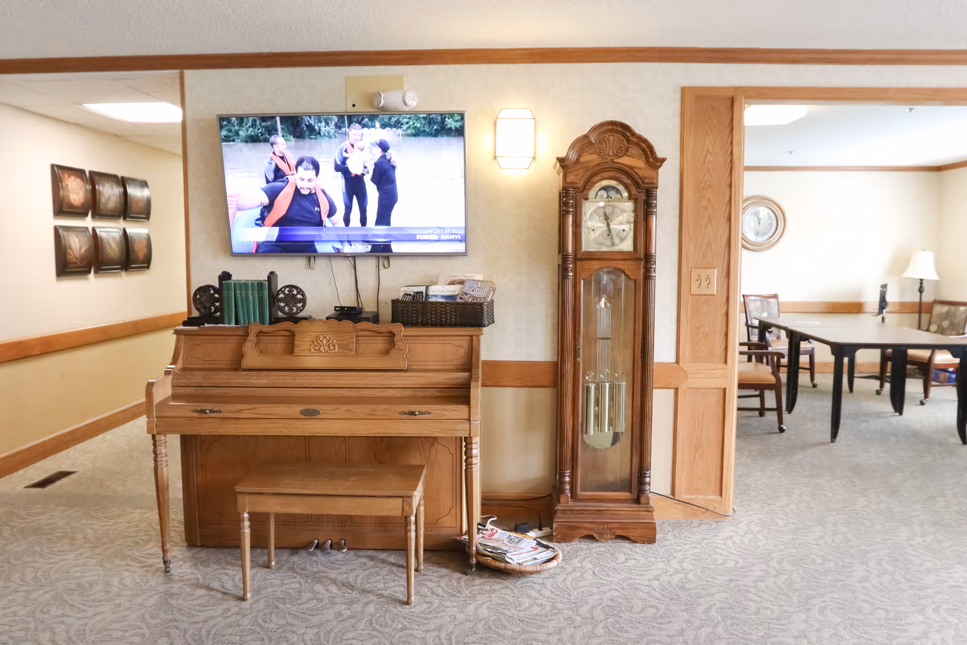 Interior view of a senior living facility room featuring a wooden piano with a matching bench, a tall grandfather clock, and a wall-mounted flat-screen TV displaying a group of people. The room has beige walls with wooden trim, carpeted flooring, and an adjacent room with a table and chairs visible through an open doorway.