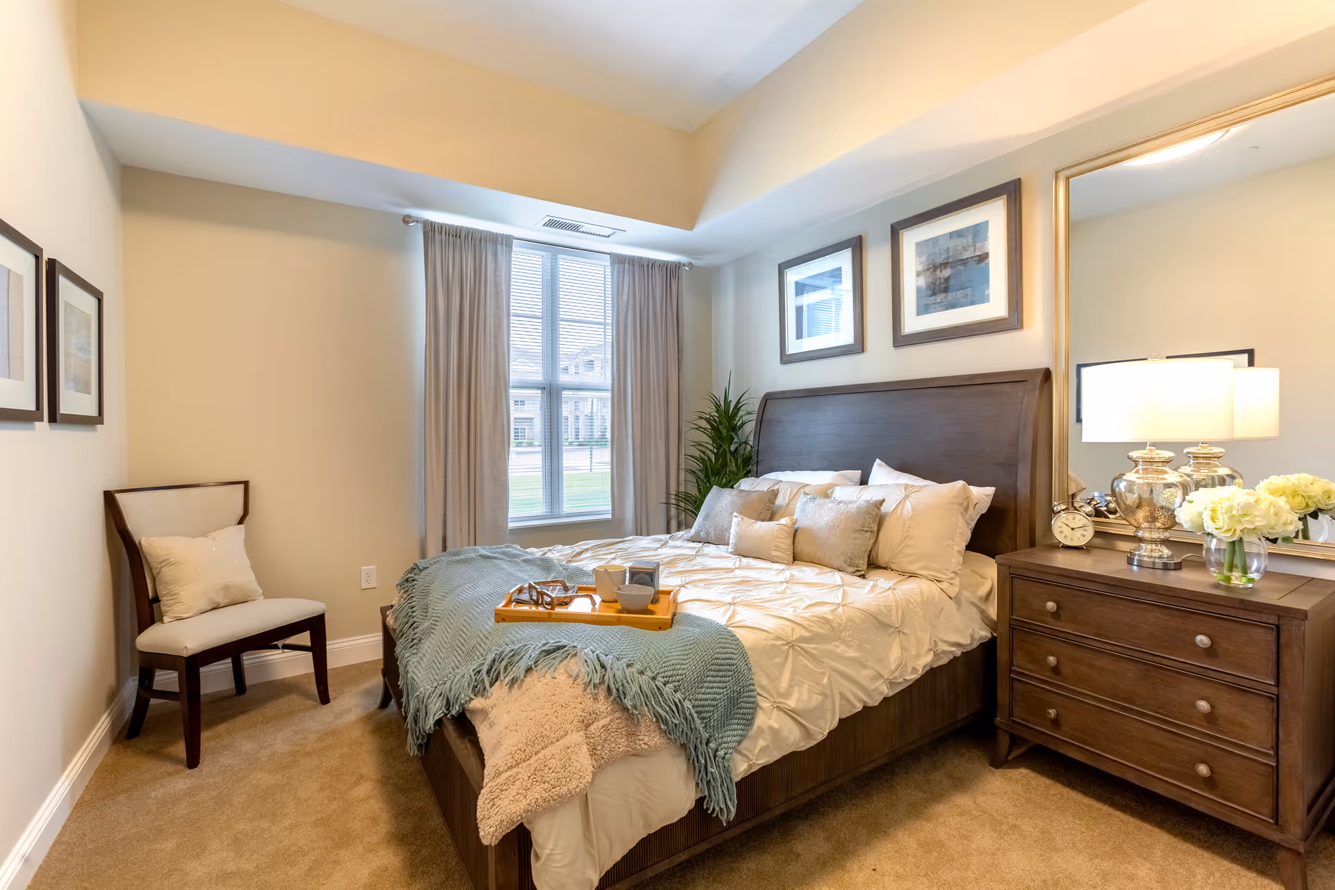A cozy bedroom with a large wooden bed adorned with multiple pillows and a light blue throw blanket. A wooden nightstand with a lamp, clock, and vase of flowers sits beside the bed. There is a chair with a cushion in the corner, two framed pictures on the wall, and a window with curtains letting in natural light.