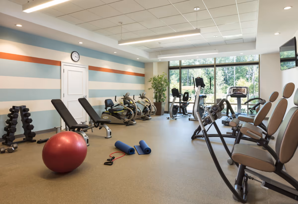 A bright fitness room with exercise machines, benches, dumbbells, a red exercise ball and yoga mats in front of large windows overlooking greenery.