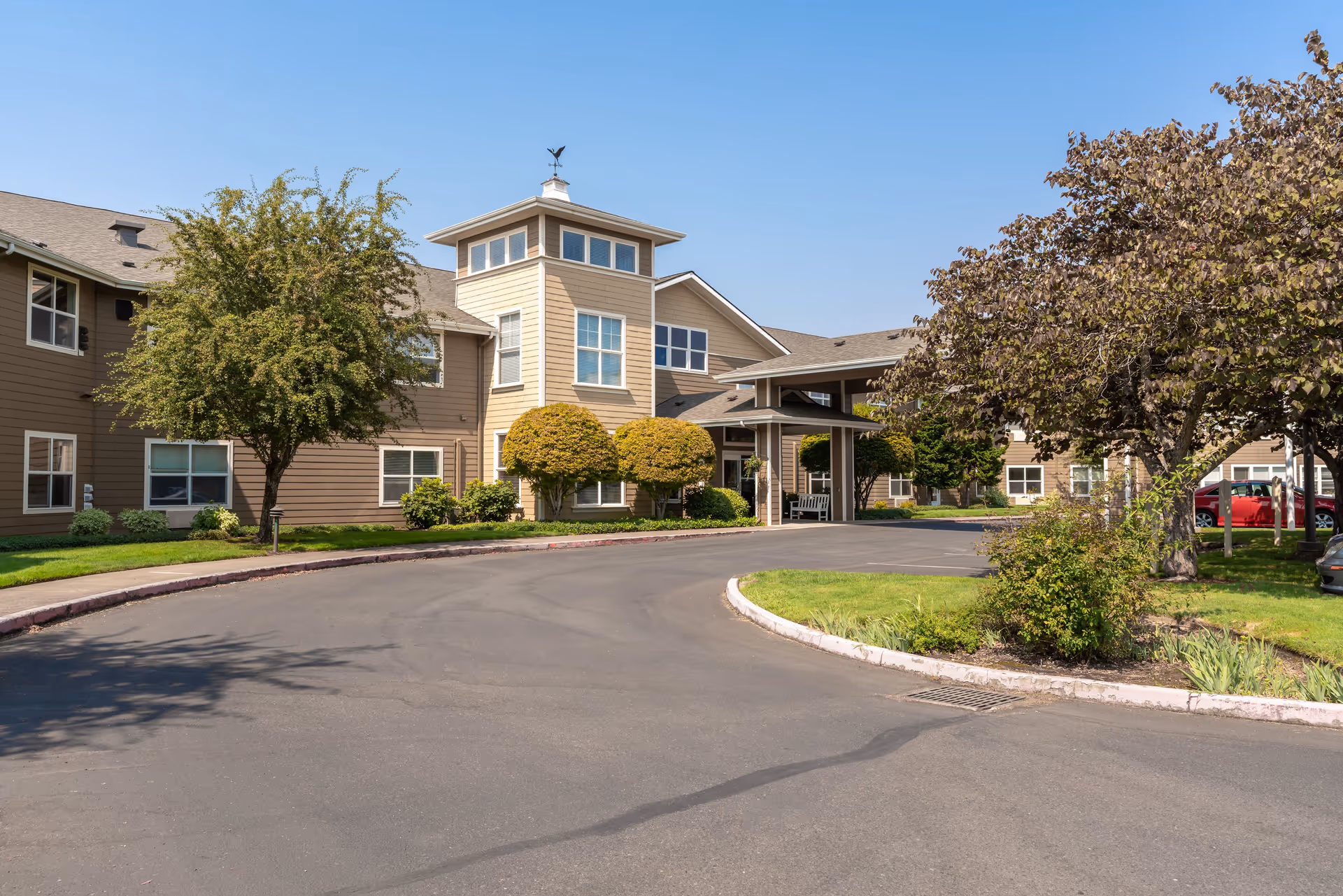 Exterior view of a senior living facility building with beige siding, multiple windows, a covered entrance, and landscaped greenery including trees and bushes under a clear blue sky.