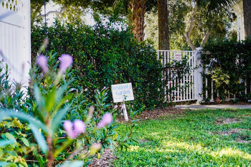 A small resident garden with flowering plants, a 'Resident's Garden' sign, and a white gate by hedges.