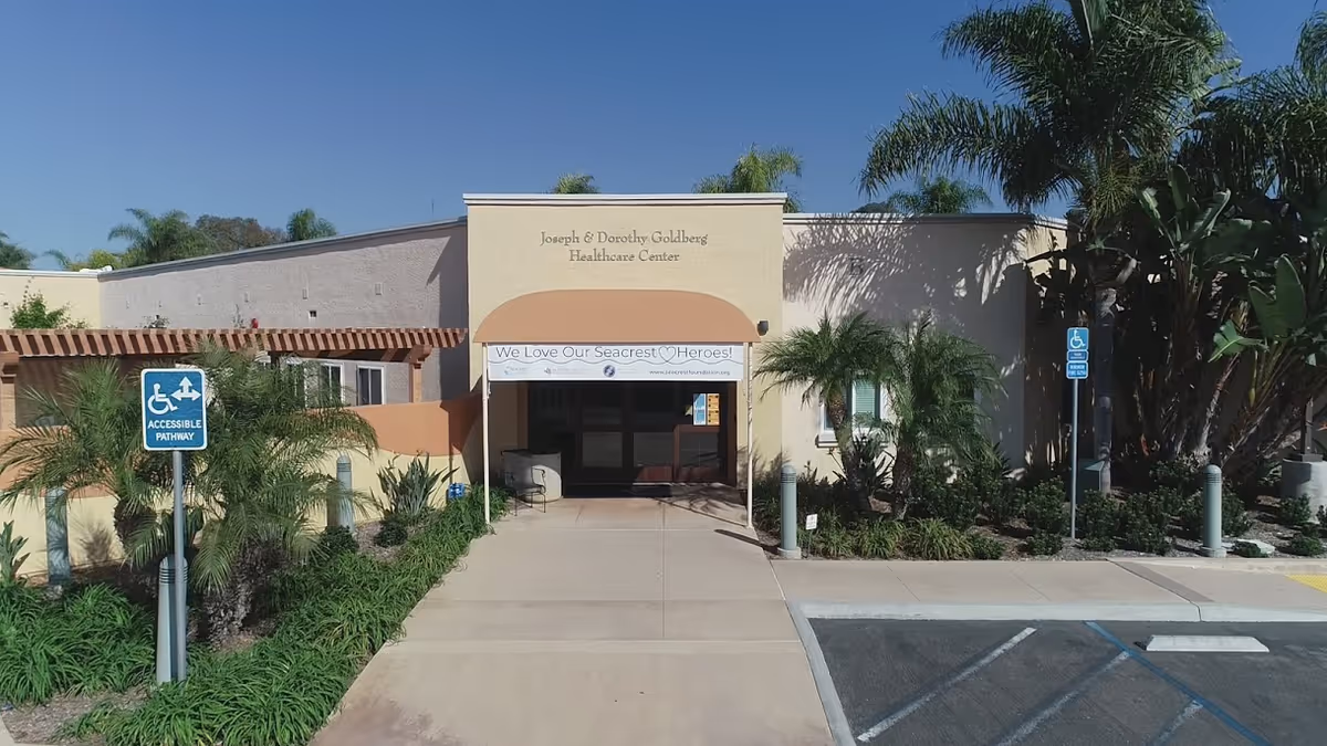 Exterior view of the Joseph & Dorothy Goldberg Healthcare Center at Seacrest Village Retirement Communities, showing the entrance with a banner that reads 'We Love Our Seacrest Heroes!'. The building is surrounded by palm trees and greenery, with accessible pathway signs visible near the sidewalk.