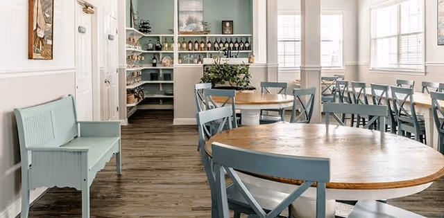 A bright dining room with round wooden tables and blue chairs arranged neatly. There is a light blue bench on the left side and shelves with decorative items and plants in the background. Large windows on the right side allow natural light to fill the room.
