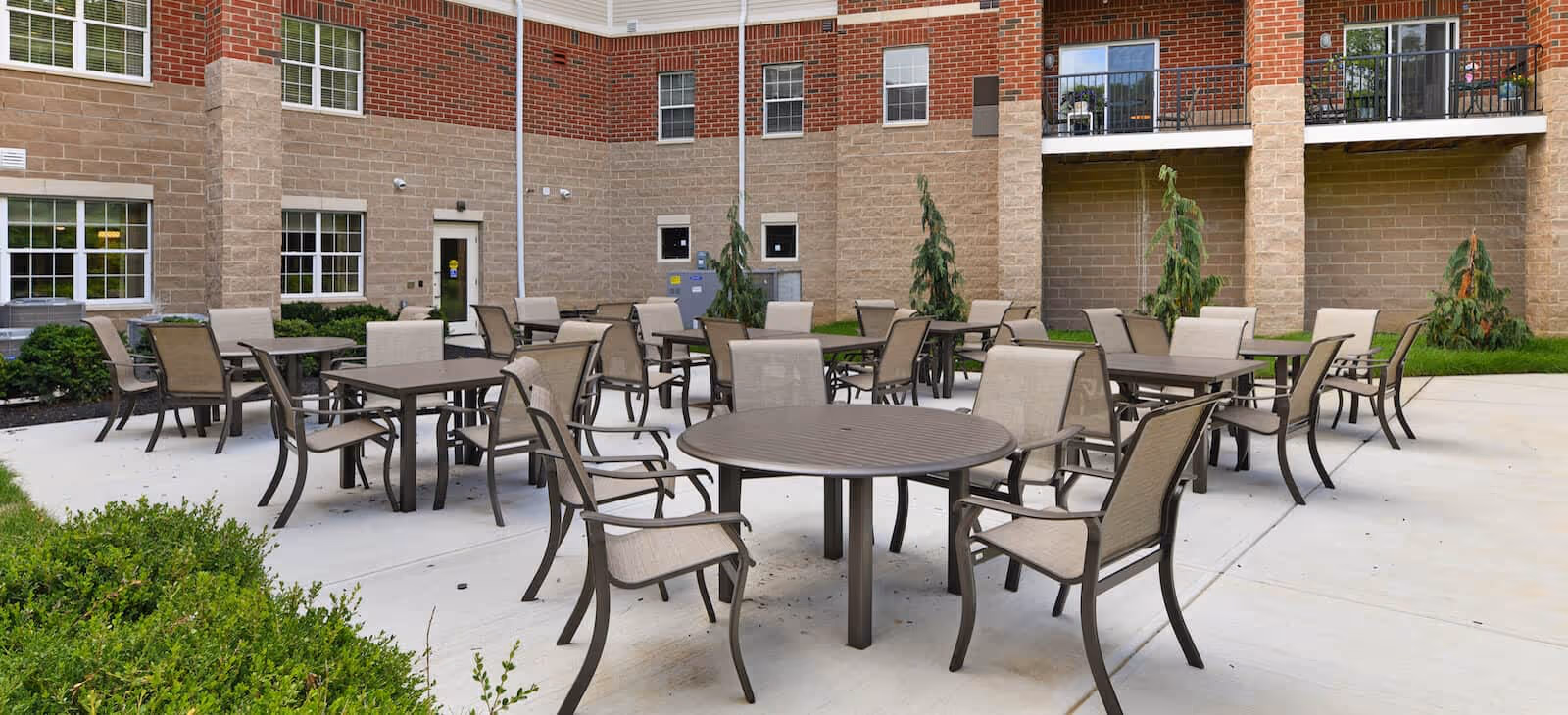 Outdoor patio area with multiple round and square tables surrounded by beige cushioned chairs, set on a concrete surface next to a brick and stone building with windows and balconies.