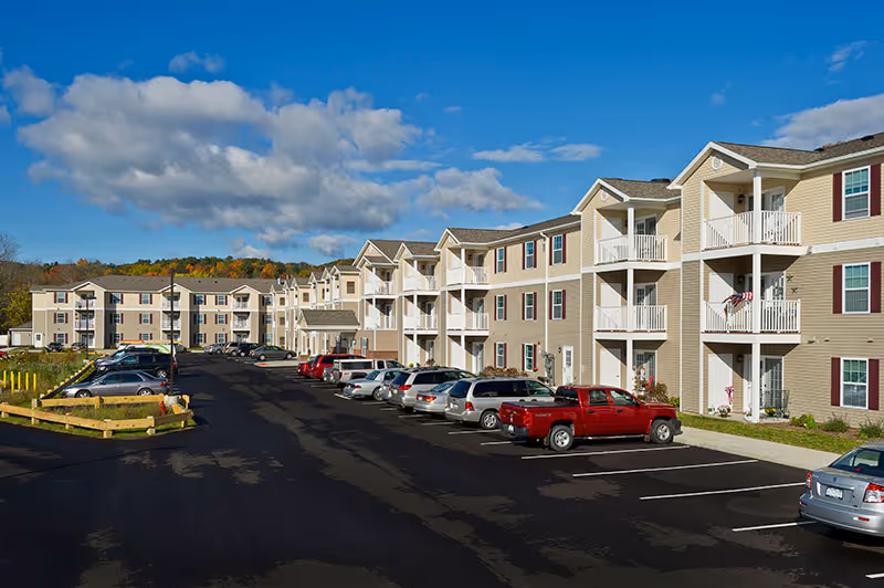 Exterior view of a multi-story beige apartment building with white balconies and maroon shutters, a parking lot with several parked cars, and a partly cloudy blue sky.