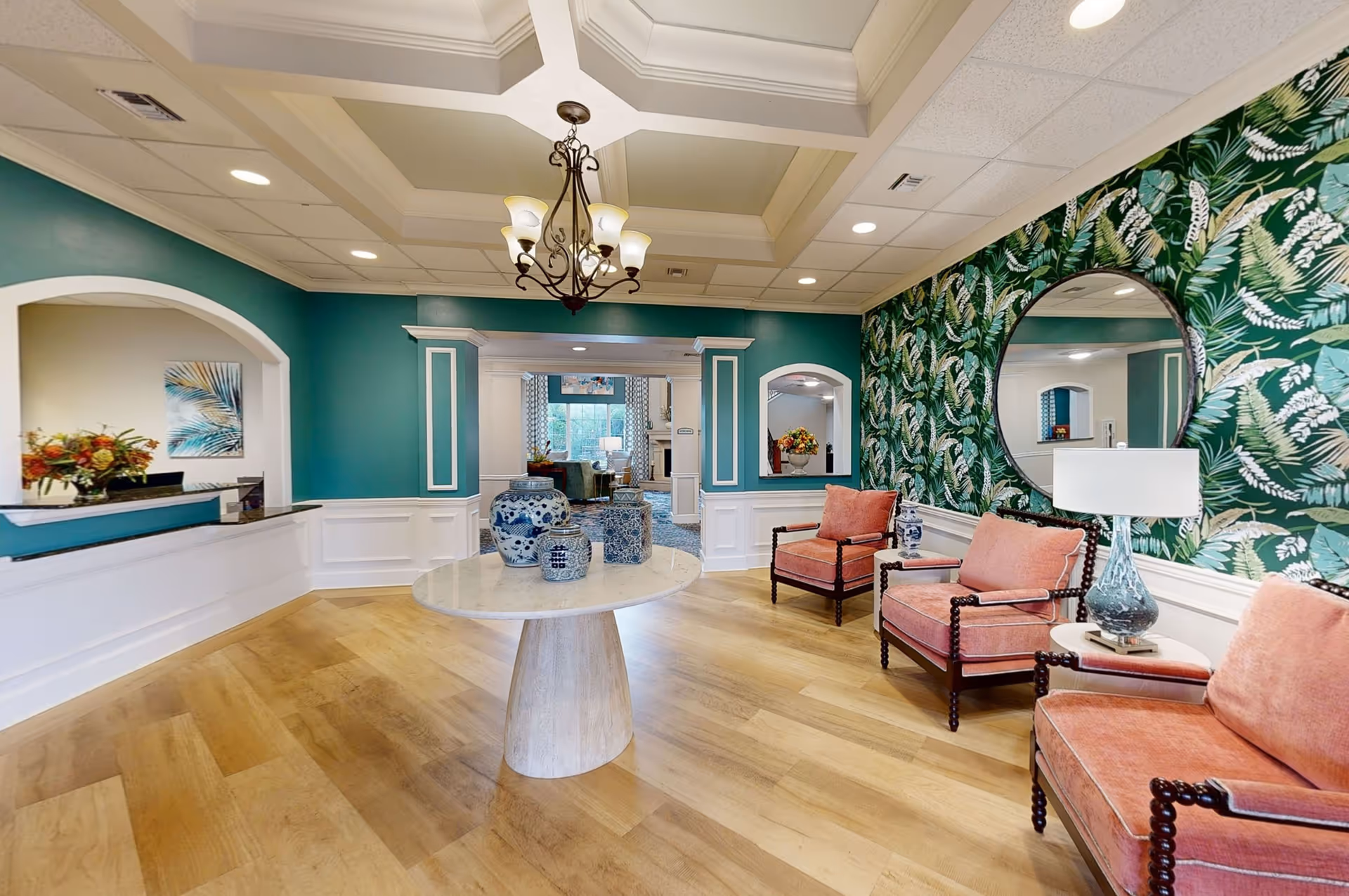 Bright lobby seating area with coral armchairs, a round table holding blue-and-white vases, tropical leaf wallpaper, and a reception desk.