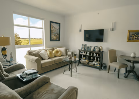 A bright living room with a large window showing an outdoor view. The room features a beige sofa with decorative pillows, two armchairs, a round glass coffee table, a TV mounted on the wall above a black cabinet filled with decorative items and framed photos. There is a small round table with a chair on the right side and framed artwork on the walls.