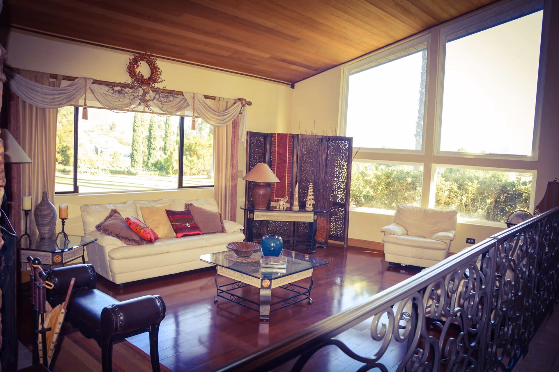 A cozy living room with large windows letting in natural light. The room features a white leather sofa with multiple decorative pillows, a matching armchair, a glass coffee table with decorative items, and a wooden screen with intricate carvings. The floor is polished wood, and the ceiling has wooden paneling. There are also curtains and a decorative wreath above the window.