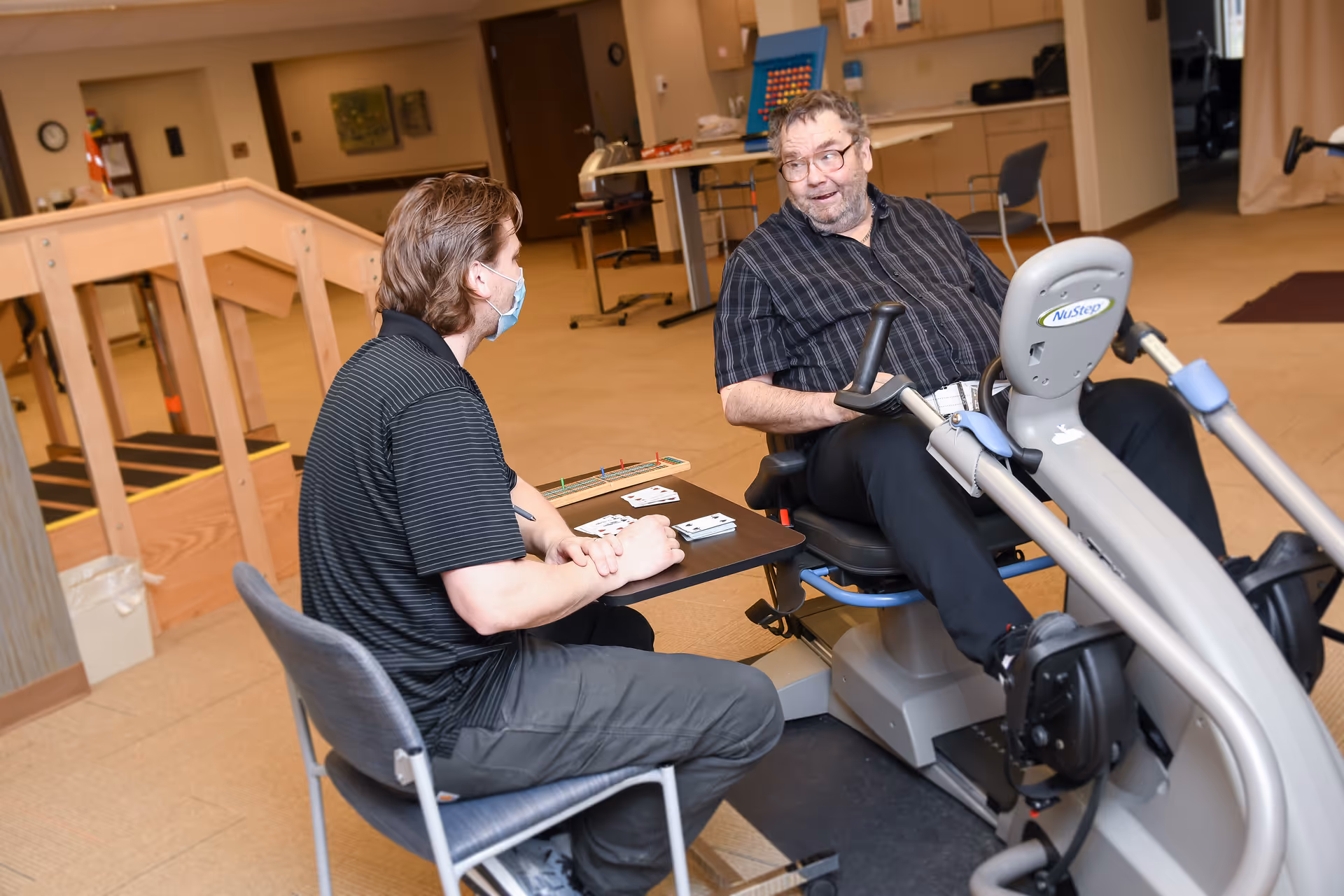 Two men in a senior care facility room; one man is seated on a recumbent exercise bike while the other man, wearing a face mask, sits on a chair across from him at a small table with playing cards and a cribbage board. The room has beige carpet, wooden handrails, and various chairs and tables in the background.