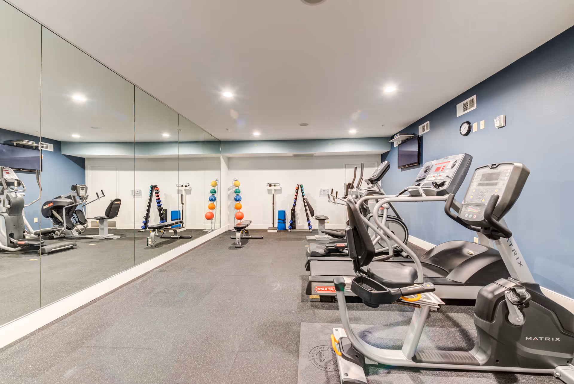 A fitness room with exercise equipment including treadmills, a recumbent bike, weight benches, dumbbells, and colorful exercise balls. One wall is covered with large mirrors, and the walls are painted blue and white. There is a wall-mounted TV and a clock on the blue wall.