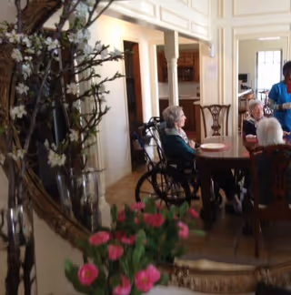 A group of elderly people sitting around a dining table in a residential care facility. One person is in a wheelchair. The room has classic decor with wooden chairs and a large mirror reflecting part of the scene. There are flowers in the foreground.