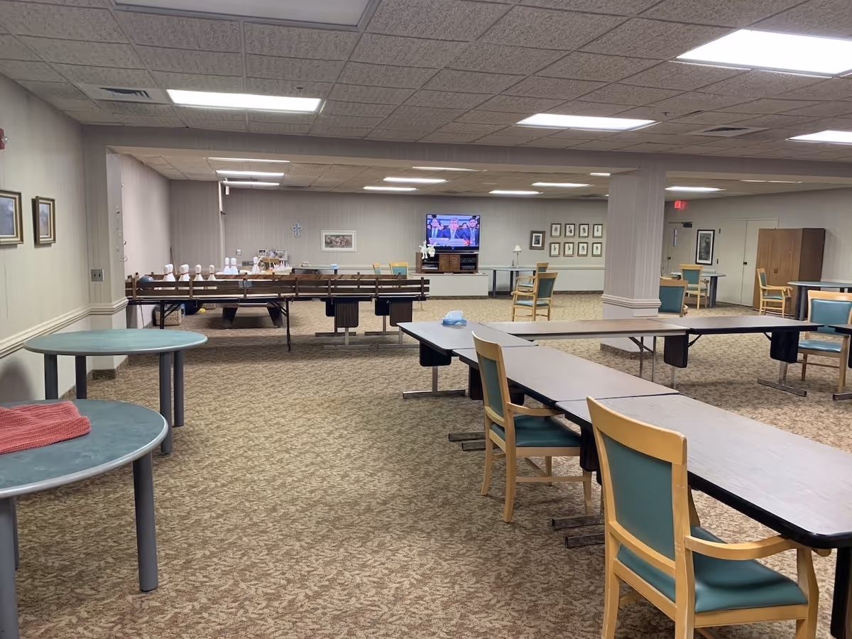 A large, carpeted common room with multiple tables and chairs arranged for group activities. There is a television mounted on the far wall showing a news broadcast, and a bowling game setup with pins on a table. The room has beige walls, framed pictures, and fluorescent ceiling lights.