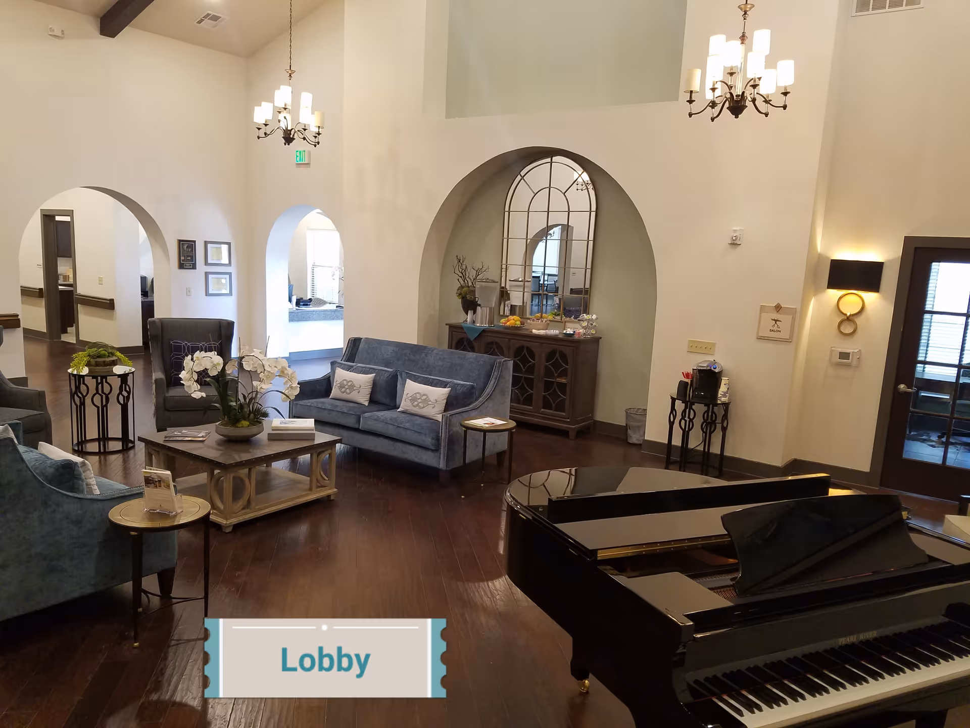 A spacious senior living facility lobby with wooden floors, blue upholstered sofas and armchairs, a coffee table with a flower arrangement, a grand piano, and two chandeliers hanging from the ceiling. There are arched doorways and a decorative mirror above a wooden cabinet with refreshments.