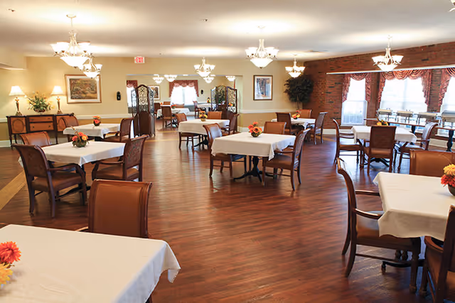 A spacious dining room with multiple tables covered in white tablecloths, each adorned with a small floral centerpiece. The room features wooden flooring, several chandeliers hanging from the ceiling, large windows with red curtains, and a brick accent wall. There are wooden chairs around the tables and decorative elements such as framed pictures and plants.