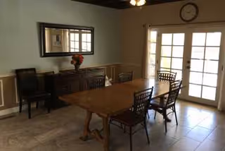 A dining room with a wooden dining table and six chairs. There is a sideboard against the wall with a vase of flowers on it, a large mirror above the sideboard, and a clock on the wall above French doors that let in natural light.