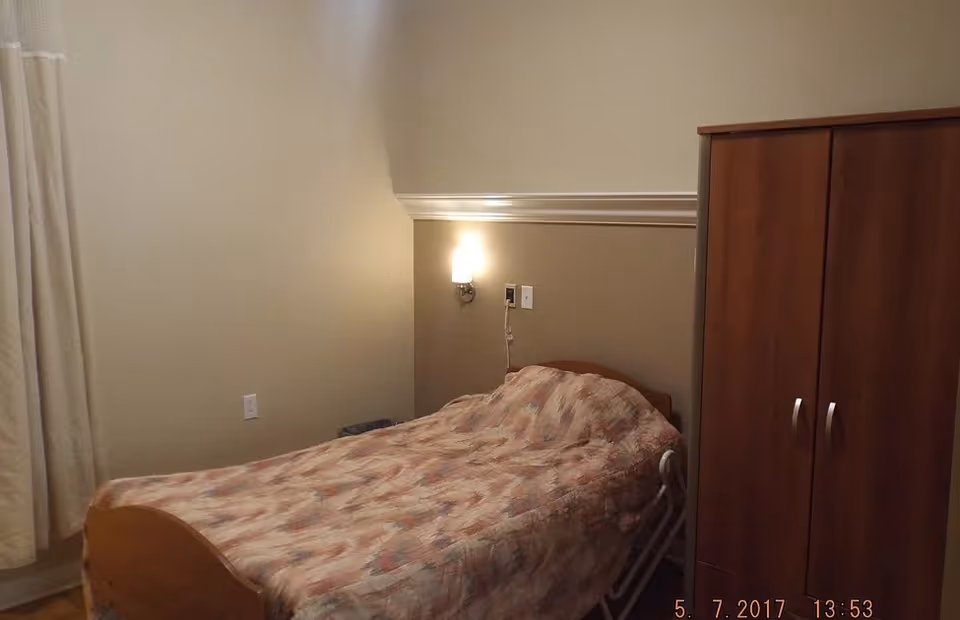 A small bedroom with a single bed covered with a patterned blanket. There is a wall-mounted light above the bed and a wooden wardrobe to the right. The walls are painted beige with a white trim, and a curtain is partially visible on the left side.