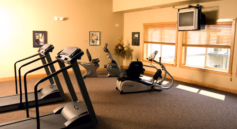 A fitness room with exercise equipment including two treadmills, two stationary bikes, and a recumbent bike. The room has beige walls, carpeted floor, framed pictures on the walls, a window with wooden blinds, and a wall-mounted television.