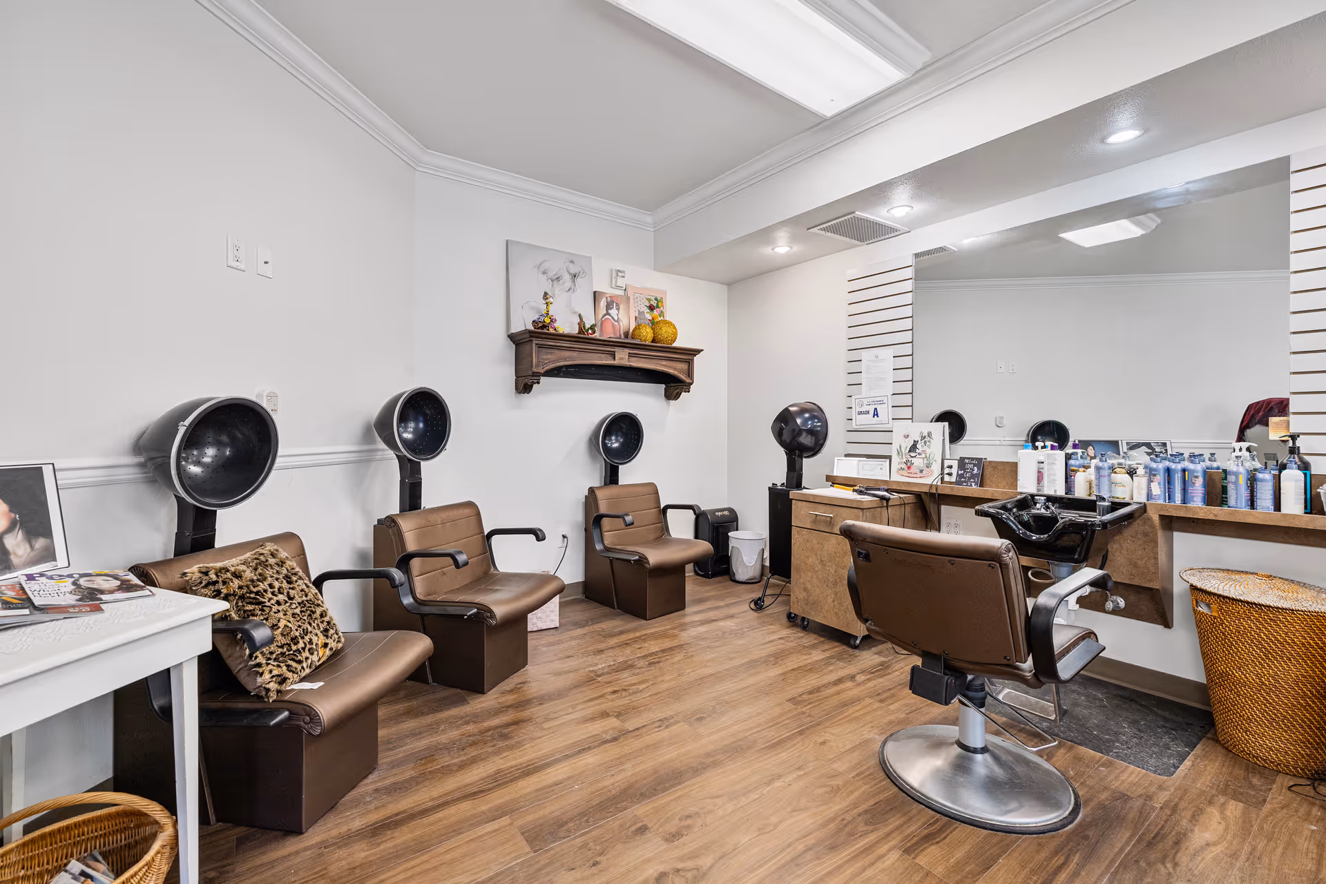 Interior view of a hair salon area with three brown salon chairs under black hair dryers along the left wall. A styling chair faces a large mirror with various hair care products on the counter. The room has wood flooring, white walls, and a small shelf with decorative items above the chairs.