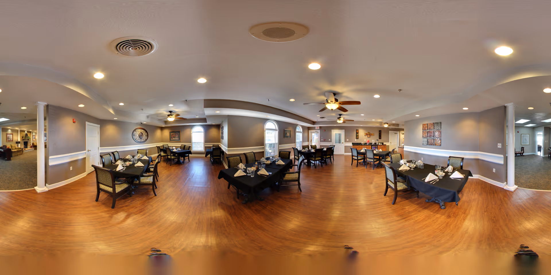 Large dining room with several tables set for meals, wood floors, ceiling fans, and soft overhead lighting.