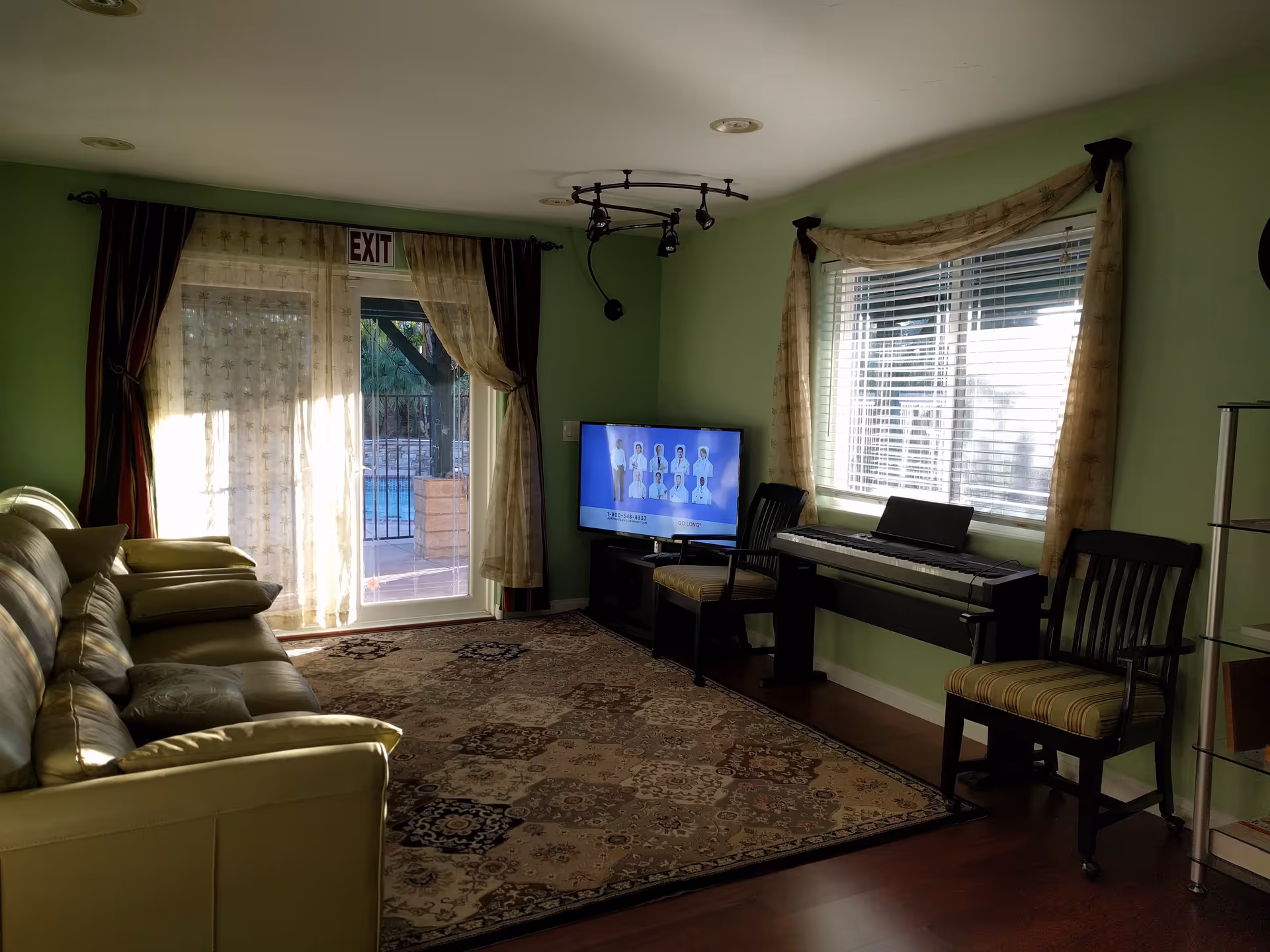 A living room with light green walls, a beige leather sofa on the left, a patterned area rug on the wooden floor, a flat-screen TV on a stand showing a commercial, two wooden chairs with striped cushions, and an electronic keyboard under a window with blinds and curtains. There is a glass door with sheer curtains leading outside to a pool area, and an exit sign above the door.