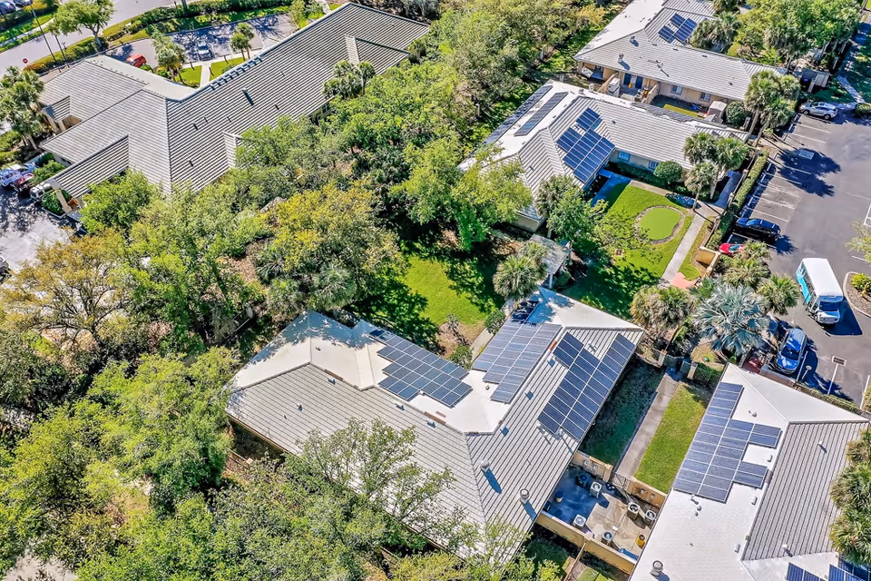 Aerial view of Healthpark Senior Living facility showing multiple buildings with solar panels on the roofs, surrounded by trees and greenery. There is a small putting green and a parking area with several cars visible.