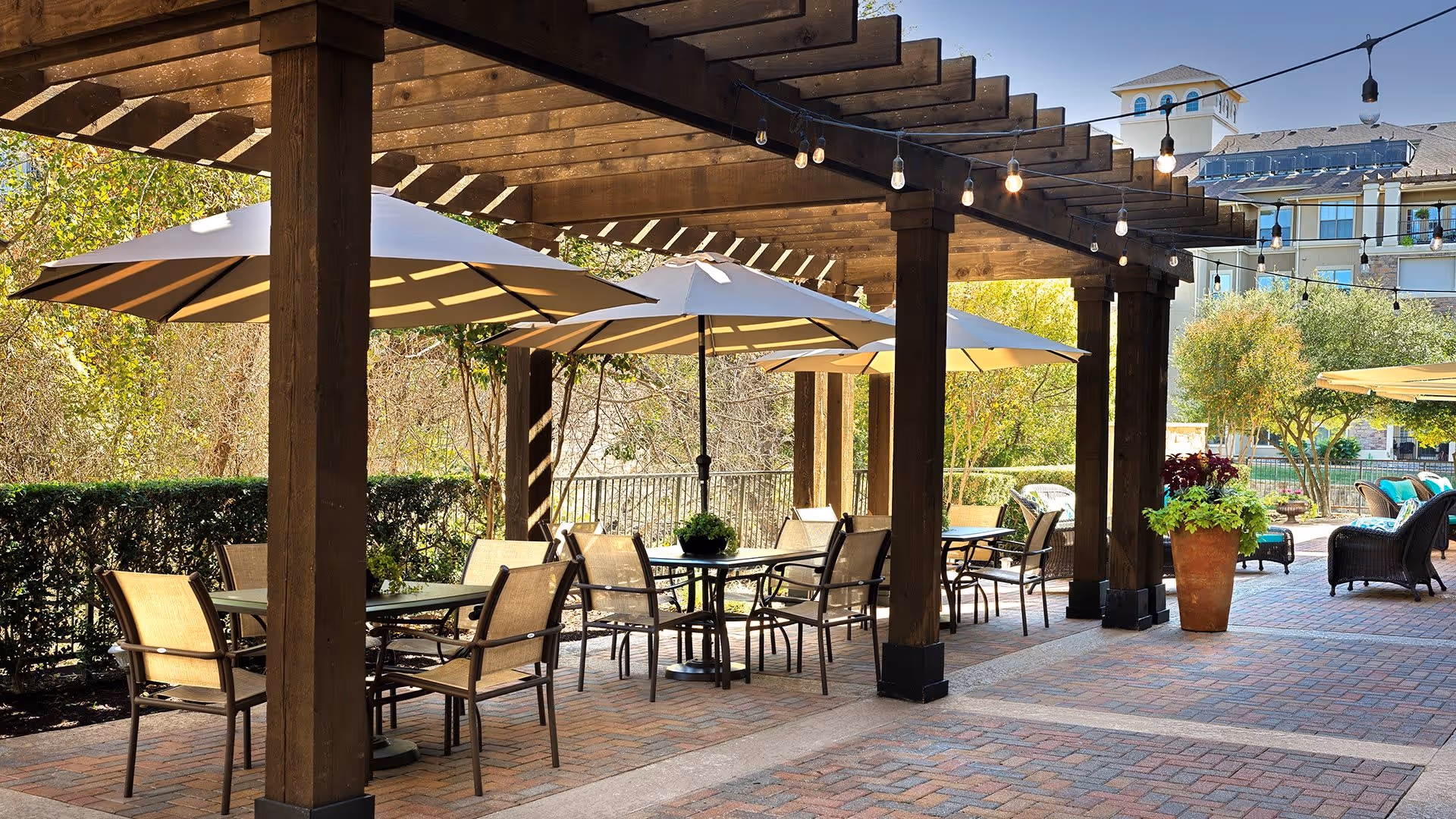 Outdoor patio area with wooden pergola, several tables with chairs, and large umbrellas providing shade. String lights hang from the pergola. There are plants and trees surrounding the patio, and a multi-story building is visible in the background.