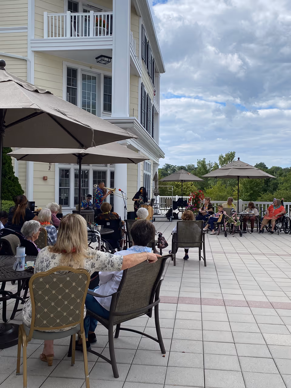 Outdoor patio area at a senior living facility with several elderly residents seated under large umbrellas, watching a live musical performance by a small band. The building is light yellow with white trim and multiple balconies. The sky is partly cloudy.