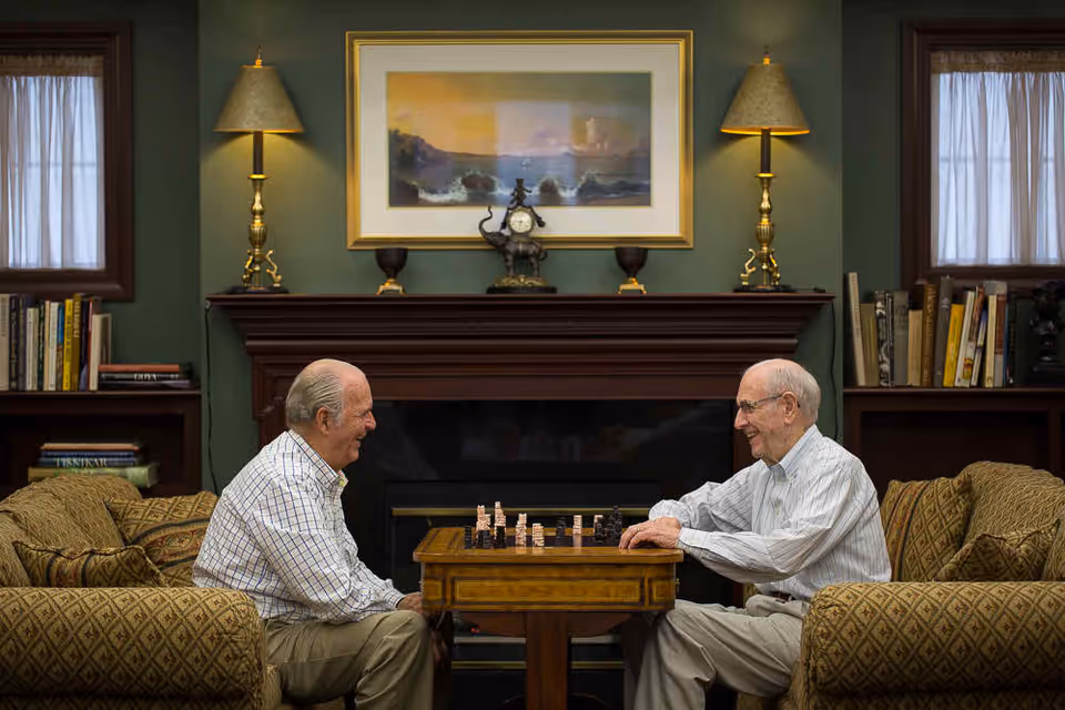 Two elderly men sitting across from each other at a small wooden table playing chess in a cozy living room with patterned armchairs, a fireplace, bookshelves, and a framed painting above the mantle.