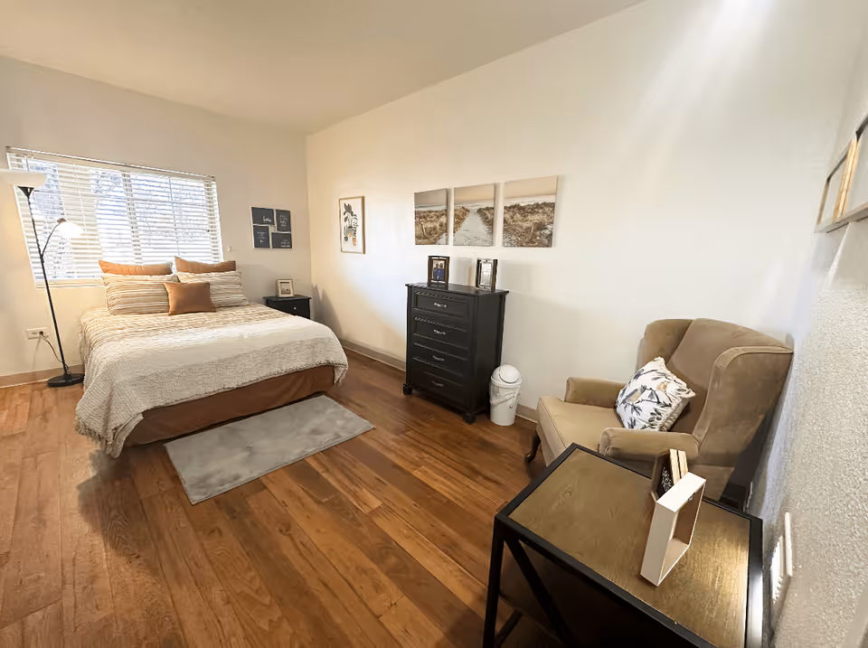 A cozy bedroom with a bed made up with beige and brown bedding, a floor lamp next to the window with blinds, a black chest of drawers with framed photos on top, a beige armchair with a decorative pillow, and a small table with a book and a decorative item. The room has wooden flooring and light-colored walls with framed artwork.