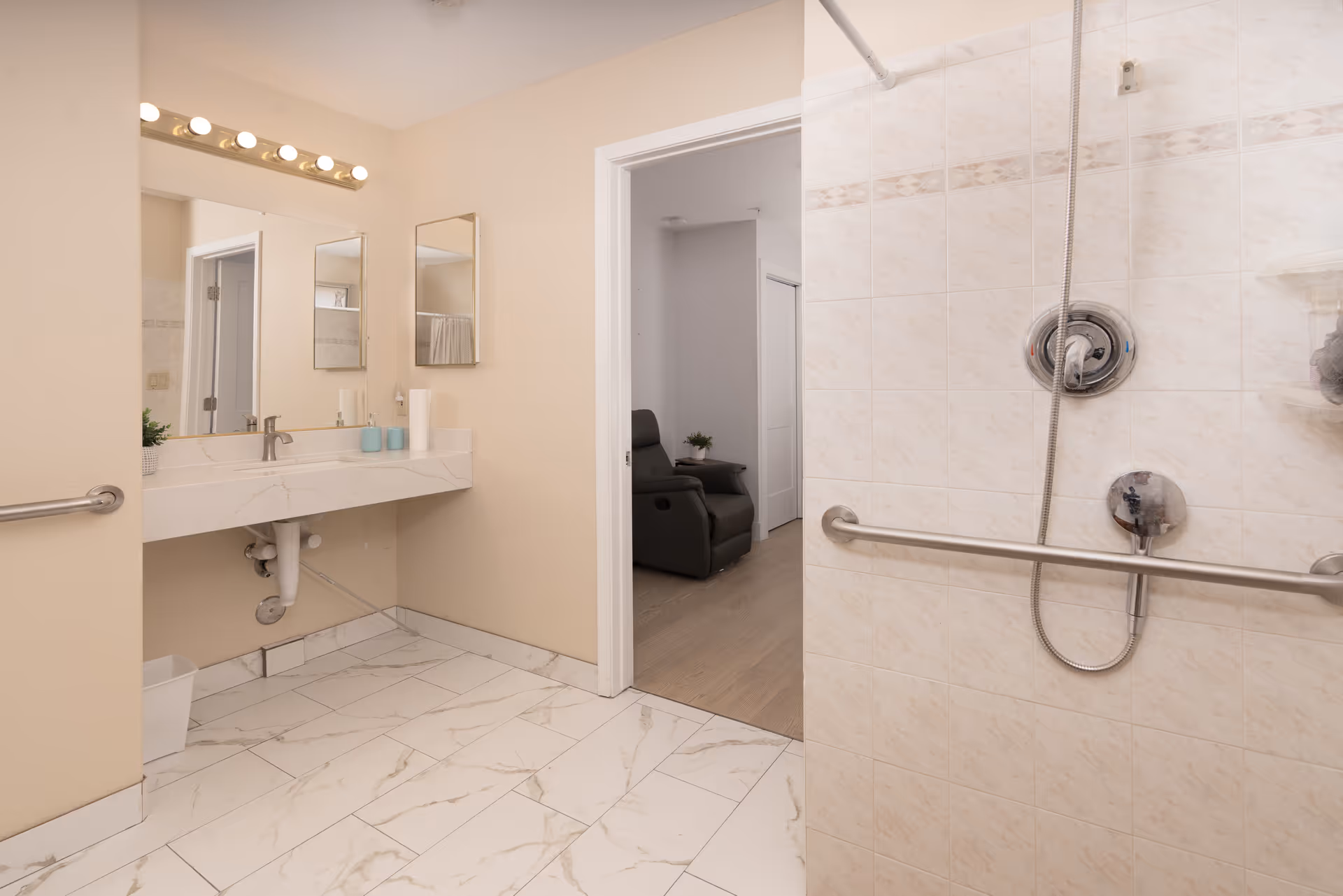 A clean and accessible bathroom with a large mirror above a white marble countertop sink. The bathroom has beige walls and white tiled floors. There is a shower area with a handheld showerhead and grab bars for safety. An open door leads to an adjacent room with a recliner chair and a small table with a plant.