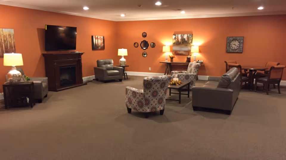 A cozy living room area with orange walls, featuring a flat-screen TV mounted above a wooden fireplace mantel, several armchairs and a sofa arranged around a coffee table, side tables with lamps, wall art including paintings and a clock, and a small dining table with chairs in the corner.
