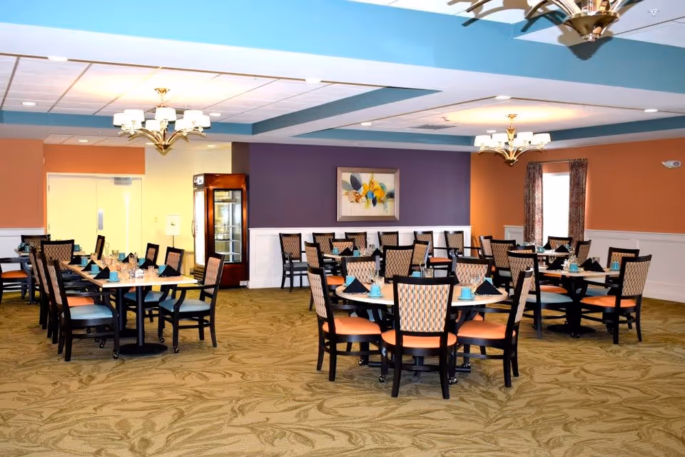 A spacious dining room with multiple round and rectangular tables set with teal cups and black napkins. The room has patterned carpet, orange and purple walls, and decorative chandeliers hanging from the ceiling. There is a framed abstract painting on the purple wall and a window with curtains on the right side.