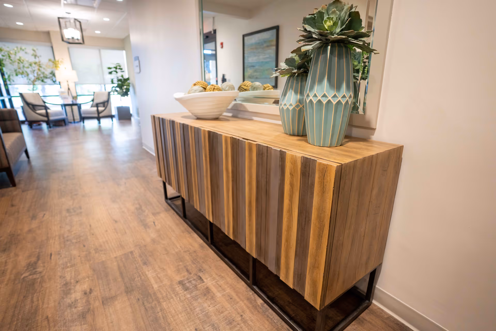 A modern wooden sideboard with vertical slats stands against a wall with a large mirror above it. On top of the sideboard are two decorative blue-green vases with plants and a white bowl filled with decorative spheres. In the background, there is a seating area with chairs, a table, plants, and large windows letting in natural light.
