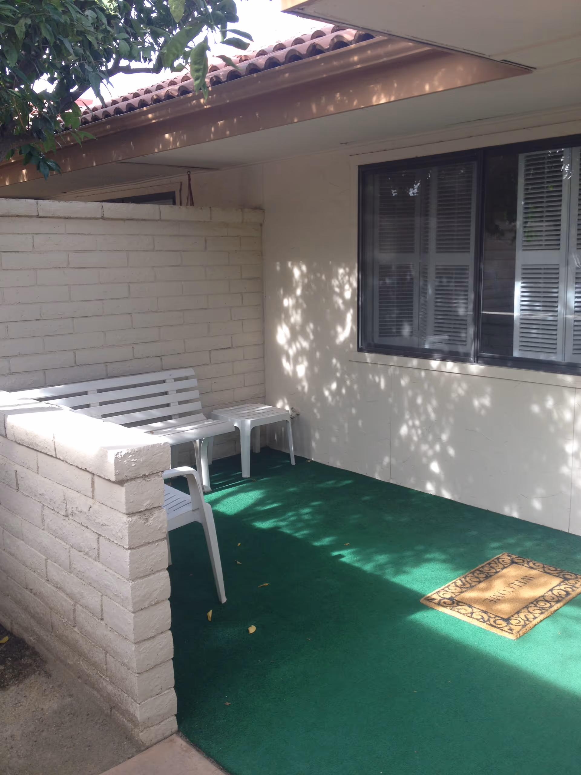 A small outdoor patio area with green outdoor carpet, a white bench, a white plastic chair, and a small white side table. The patio is enclosed by a low white brick wall and has a window with closed blinds on the adjacent building wall. A doormat is placed near the entrance, and tree shadows are cast on the walls and floor.