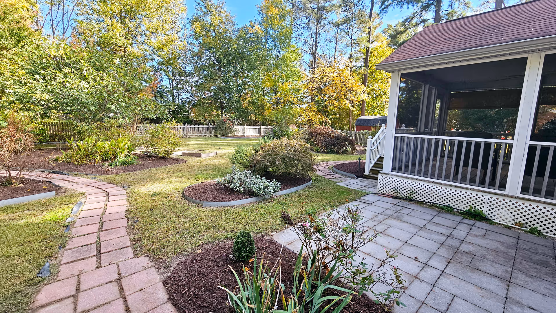 A landscaped outdoor garden area with a paved walkway curving through mulched flower beds and green grass. There is a white picket fence in the background and a screened porch attached to a building on the right side. Trees with autumn foliage surround the garden.