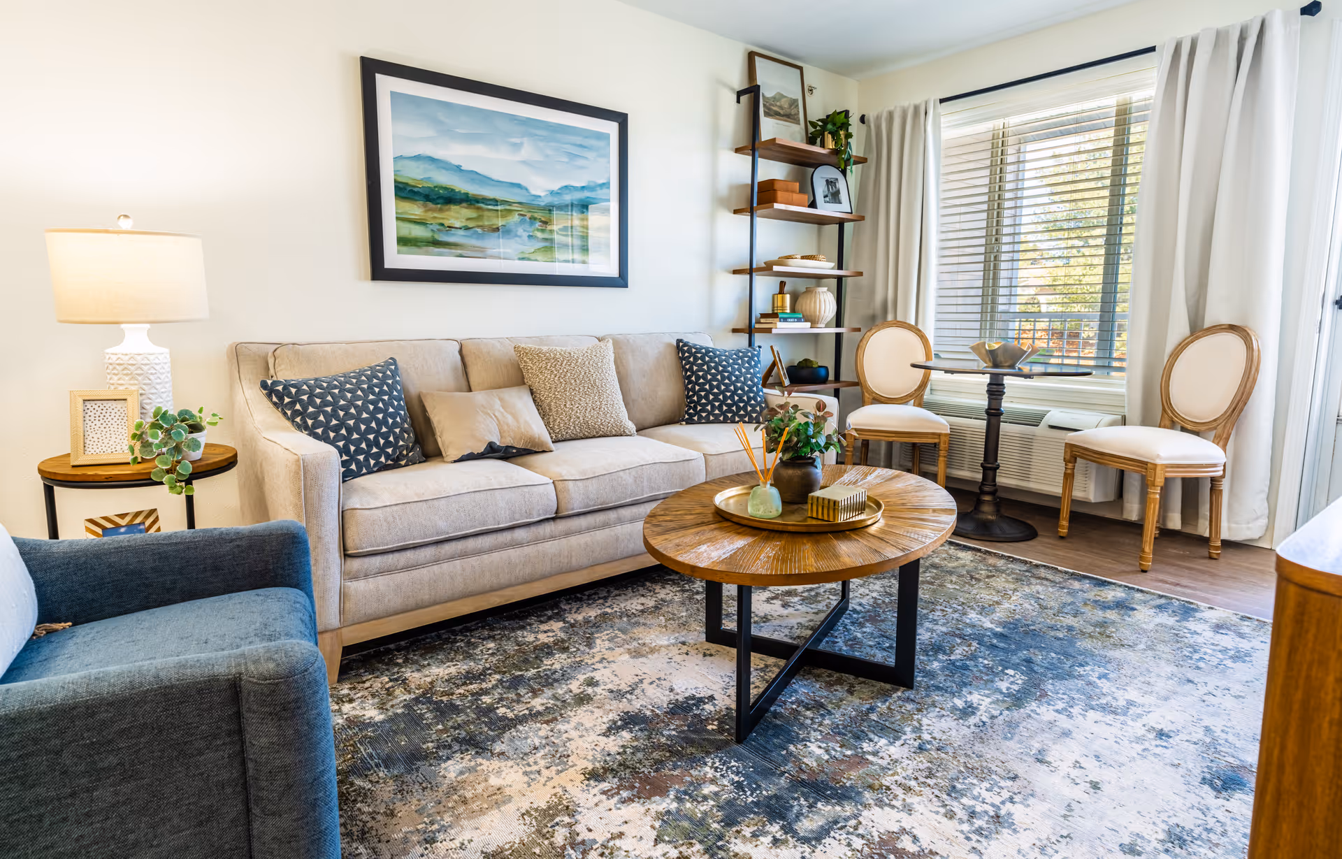 A bright and cozy living room with a beige sofa adorned with patterned cushions, a blue armchair, a round wooden coffee table with decorative items, a side table with a lamp and plant, a tall shelving unit with books and decor, and two upholstered chairs near a window with white curtains and blinds.