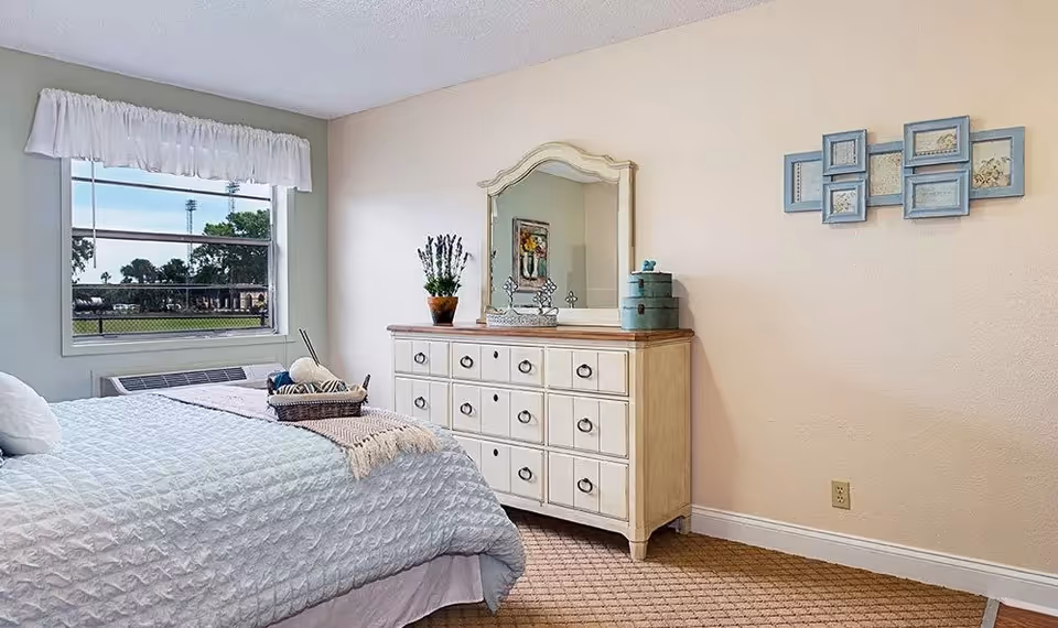 A cozy bedroom with a neatly made bed covered in a light blue quilt. There is a basket with yarn and knitting needles on the bed. A large window with a white valance lets in natural light and shows a view of trees and a field outside. Against the wall is a cream-colored dresser with multiple drawers, topped with a decorative mirror, a potted plant, and some small decorative items. A blue multi-photo frame hangs on the beige wall above an electrical outlet.