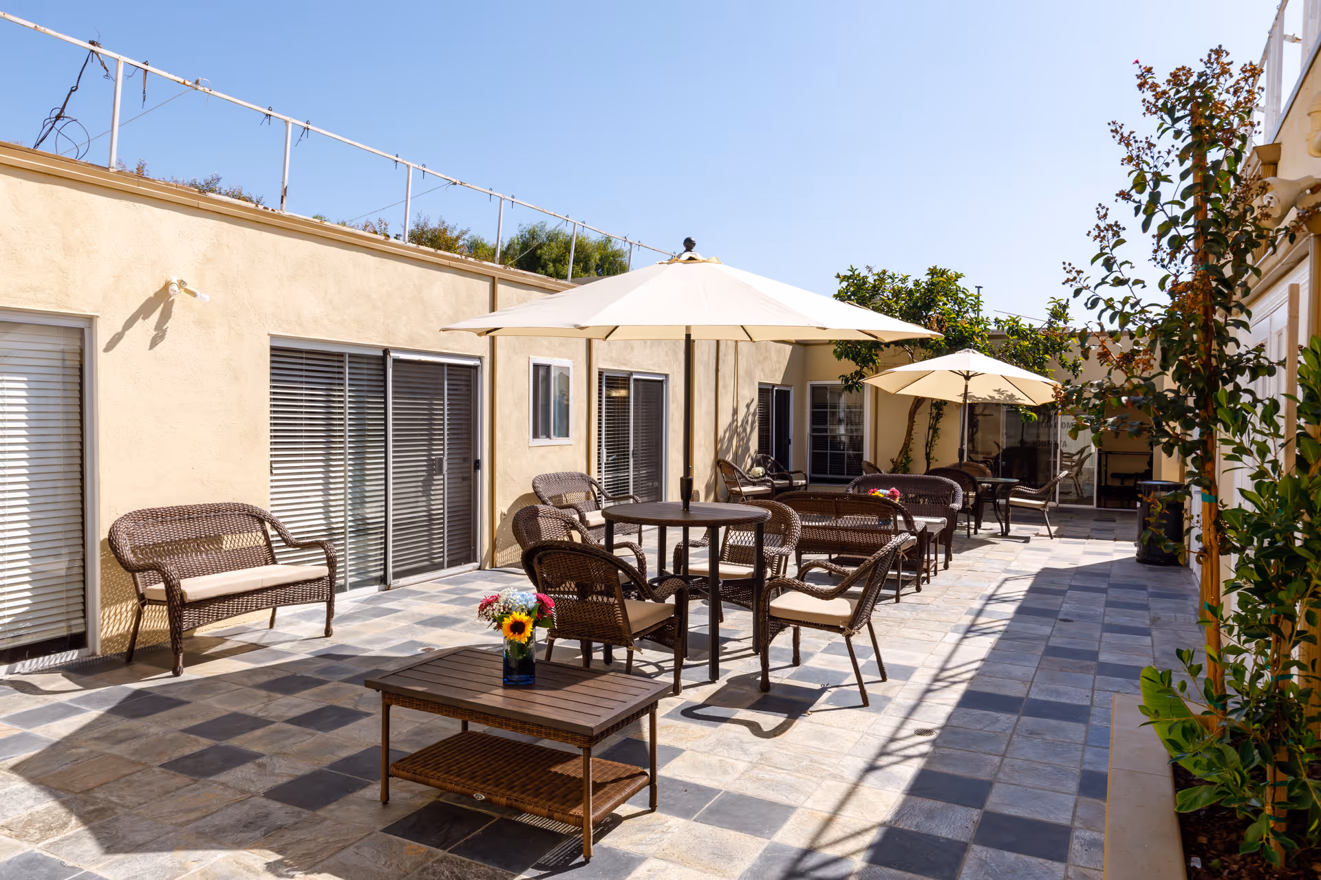 Outdoor patio area at Ocean Pointe Healthcare Center with wicker chairs and tables, some shaded by large white umbrellas. The patio is paved with stone tiles and decorated with potted flowers and plants along the edges. The building walls are beige with several windows and sliding glass doors.