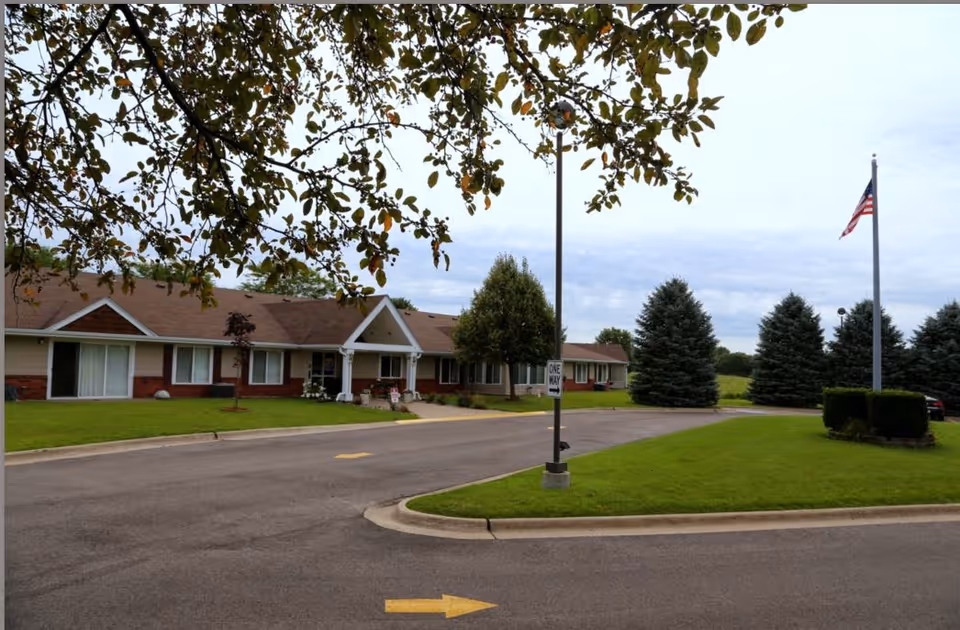 Exterior view of a single-story building with a brown roof and beige walls, surrounded by green grass and trees. There is a paved driveway with a 'One Way' sign and an American flag on a flagpole on the right side of the image. The sky is cloudy.