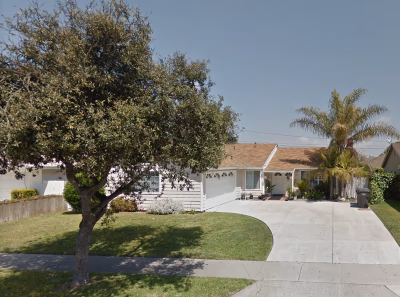 Single-story house with a light-colored exterior, a two-car garage, a driveway, and a front yard with a large tree and a palm tree under a clear blue sky.