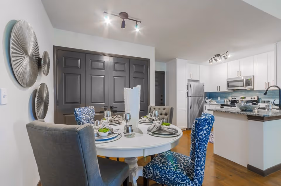 A modern dining area with a round white table set for four, featuring gray and blue patterned chairs. The background shows a kitchen with white cabinets, stainless steel appliances, and a blue tile backsplash. Decorative metallic wall art is mounted on the left wall.