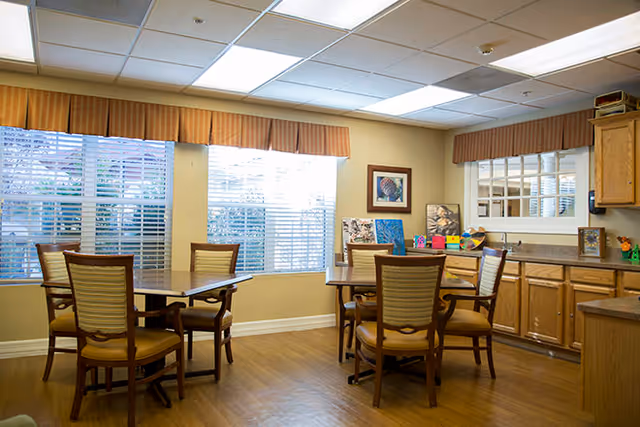 Bright communal dining/activity room with wooden tables and chairs, large windows with blinds, and a kitchenette with cabinets.