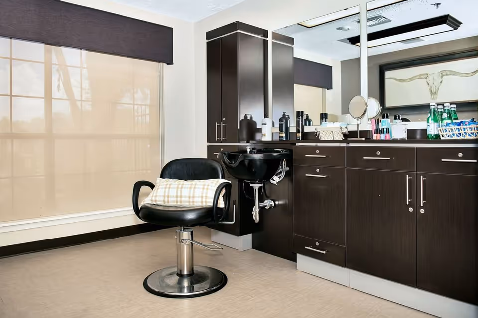 A salon area inside a nursing and rehabilitation center featuring a black salon chair with a plaid cushion, a black wash basin attached to a dark wood cabinet, various hair and beauty products on the countertop, and a large mirror reflecting the room.