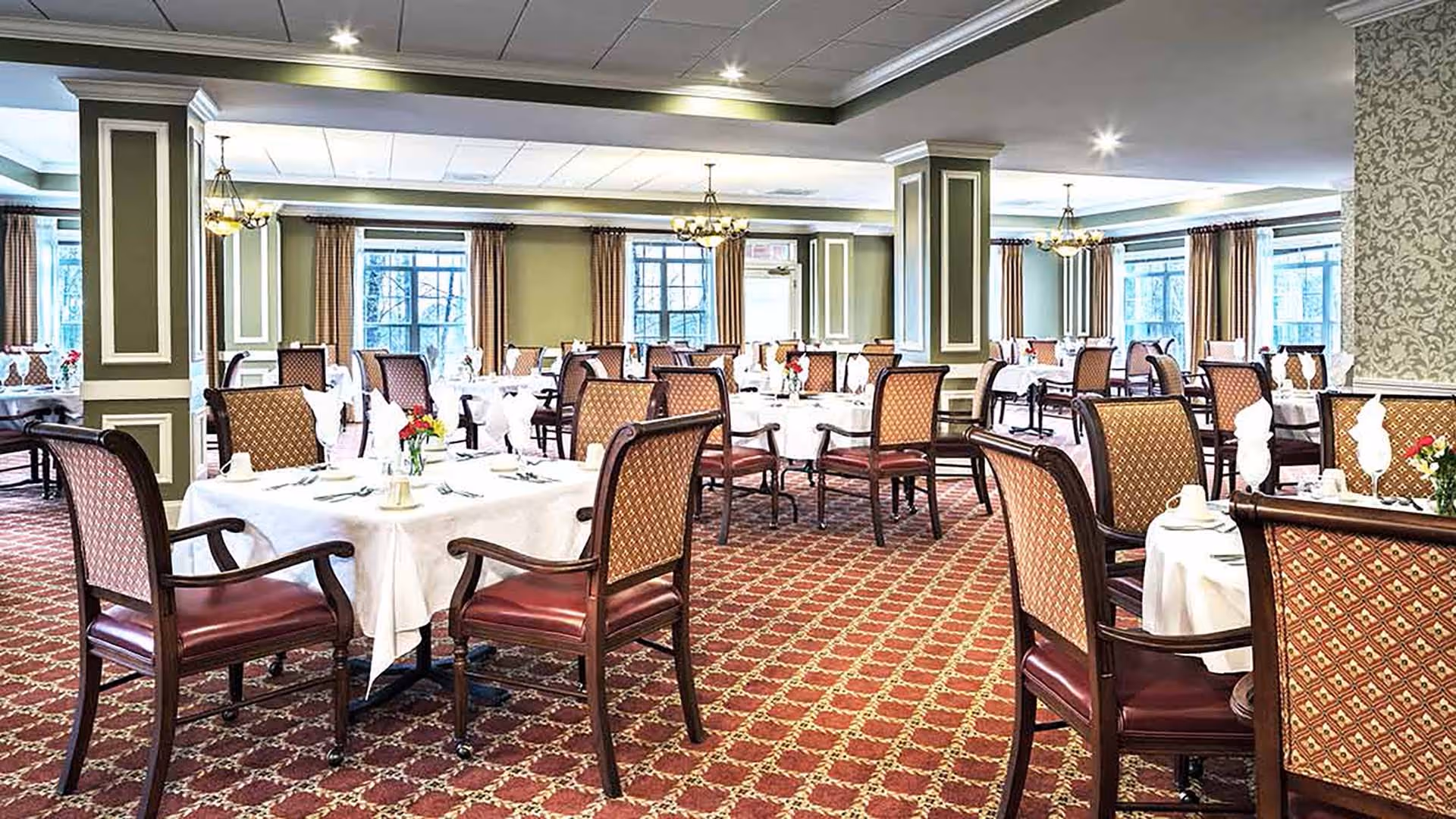 A spacious dining room with multiple tables covered in white tablecloths, each set with napkins, plates, and silverware. The room features patterned carpet, upholstered chairs with wooden frames, large windows with curtains, and decorative columns. Chandeliers hang from the ceiling providing warm lighting.
