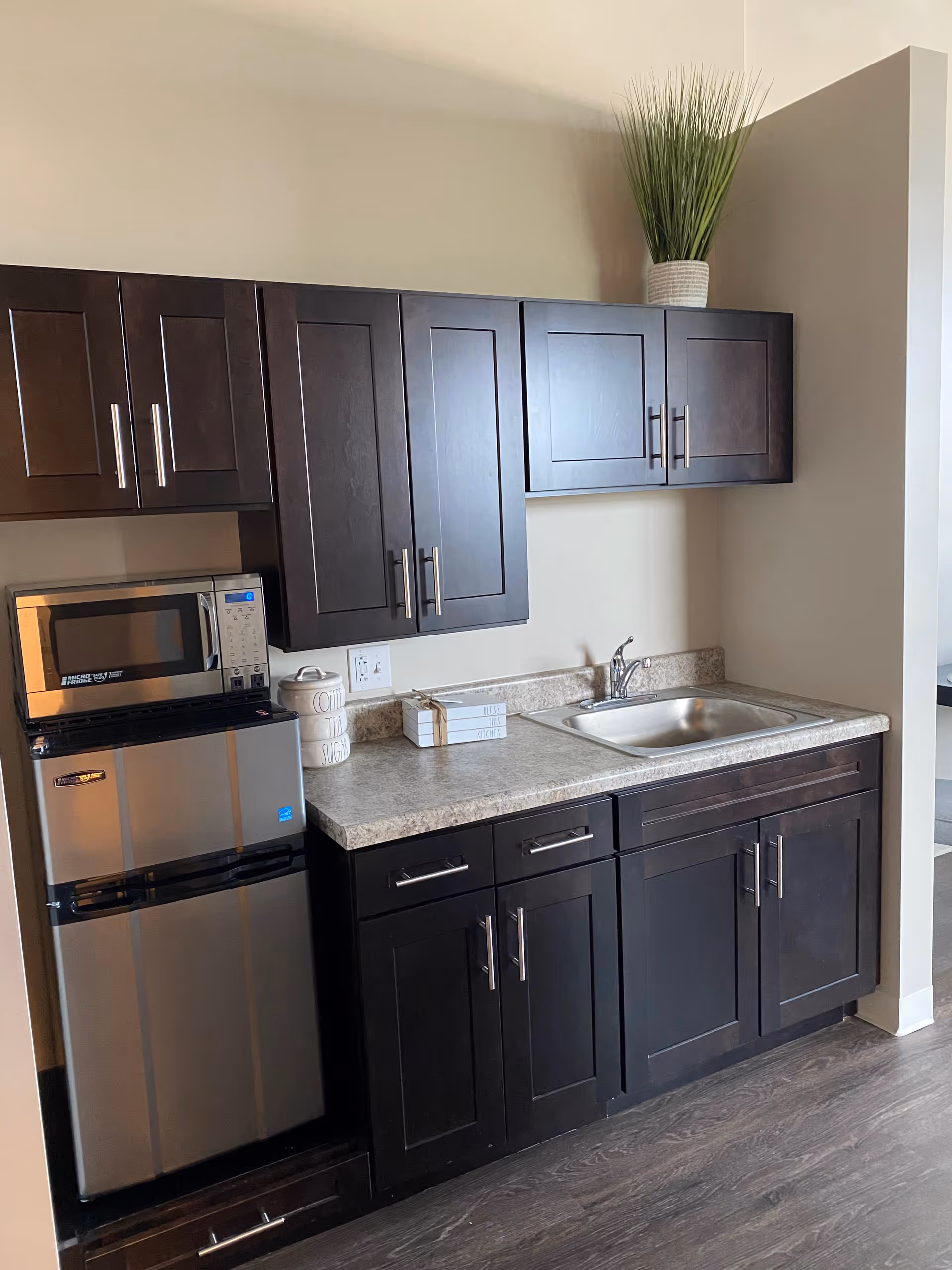 A small kitchen area with dark wooden cabinets, a stainless steel mini refrigerator, a microwave on top of the refrigerator, a countertop with a sink, decorative containers labeled coffee, tea, and sugar, a small stack of books, and a potted plant on top of the upper cabinets.