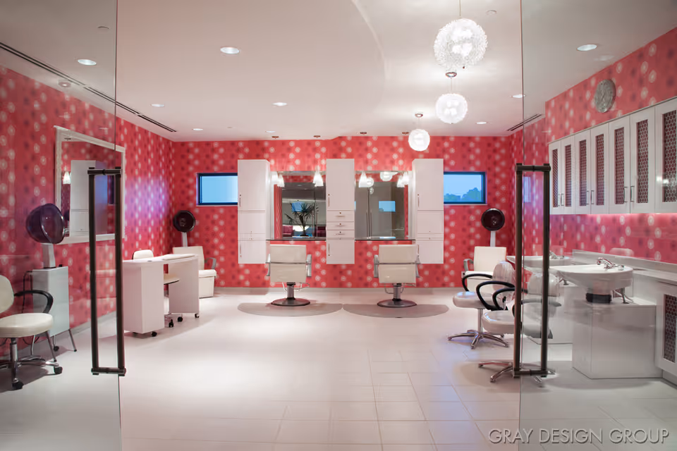 Interior view of a salon area with bright pink patterned walls, white tiled floor, and modern white salon chairs in front of mirrors. The room features glass doors, white cabinetry, hair dryers, and pendant lights hanging from the ceiling.
