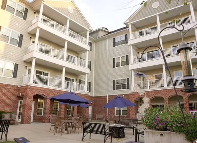 Outdoor courtyard area of a senior living facility with multiple tables and chairs under blue umbrellas, surrounded by a multi-story building with balconies and brick lower walls. There are plants and a bird feeder visible in the foreground.